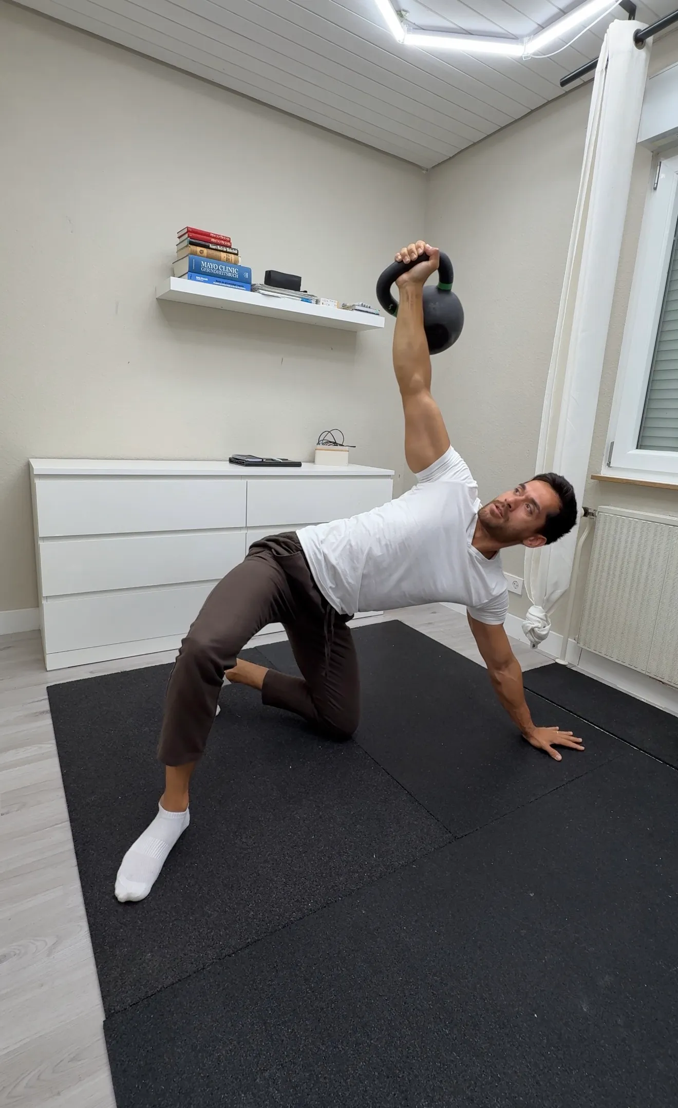 Eric Becker kniet auf einer Trainingsmatte und hält eine Kettlebell mit gestrecktem Arm über dem Kopf